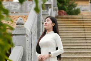 A woman in a white traditional dress stands on a stone staircase outdoors, showcasing elegance.