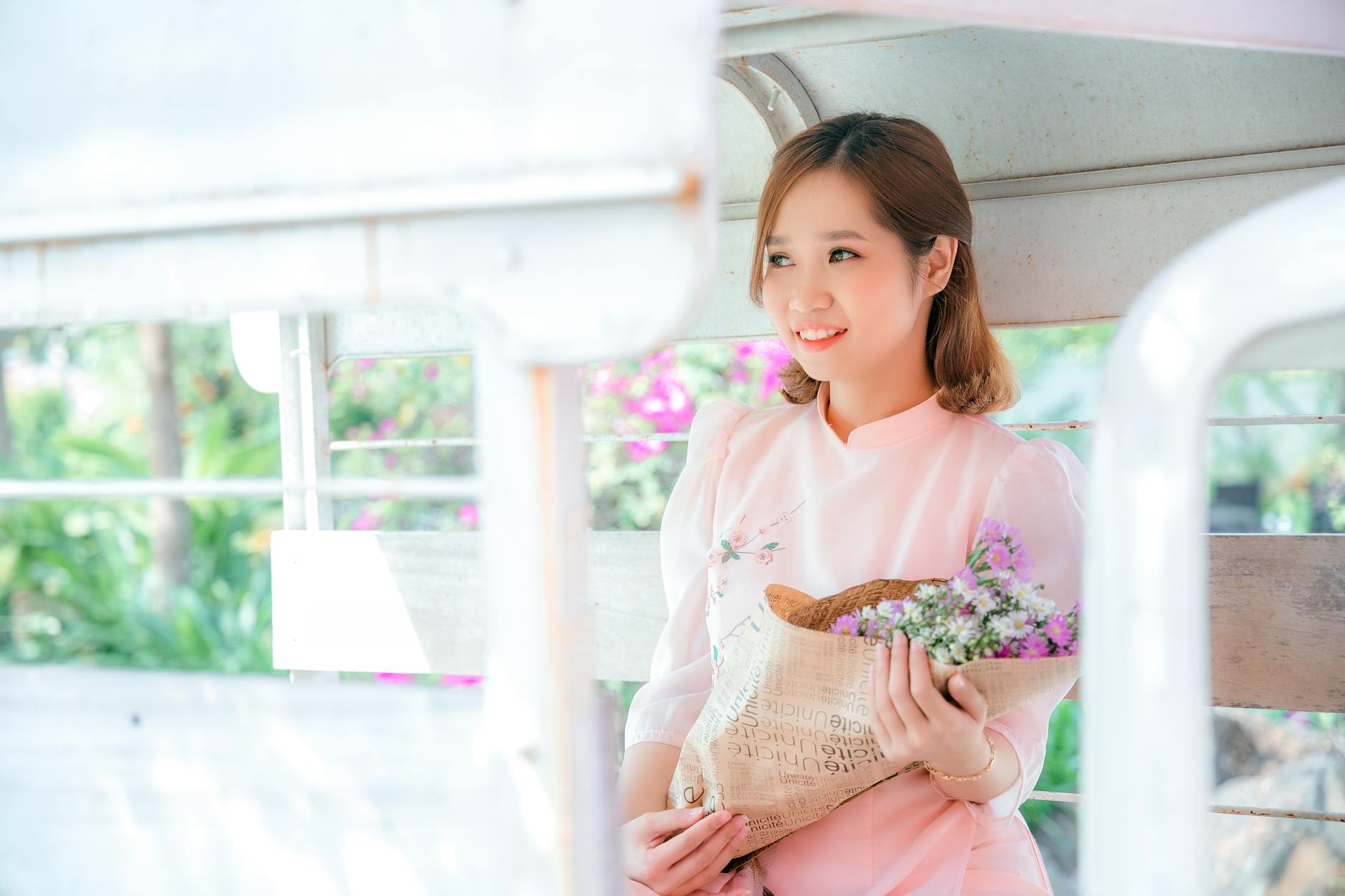 A young Asian woman in a light pink dress smiles while holding a bouquet in a sunlit setting.