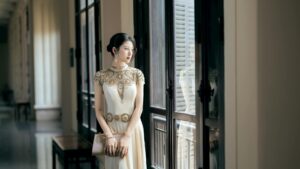 Elegant woman in a beautifully decorated gown gazing through a window indoors.