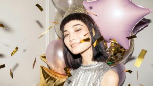 Smiling woman in a festive setting with colorful balloons and confetti.