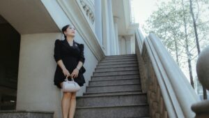 Sophisticated woman in black dress and heels standing beside an outdoor marble staircase.